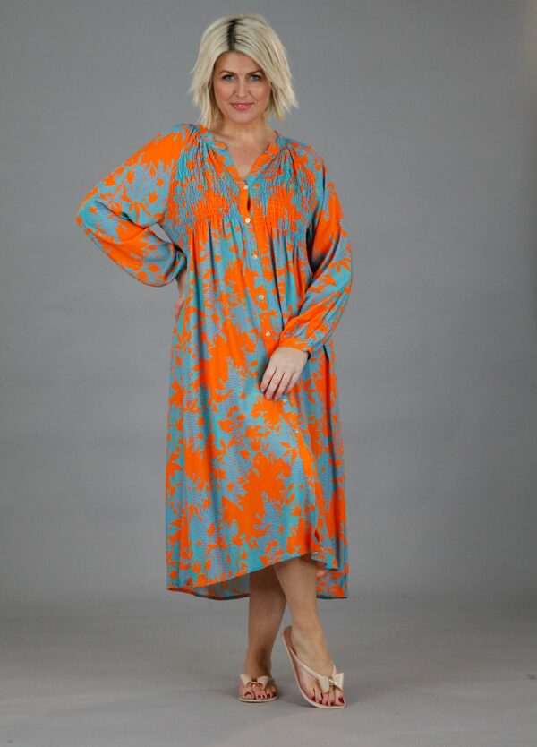 a blonde lady in an orange longsleeved, mid length dress with an abstract turquoise print, wearing silver sandals against a plain grey background
