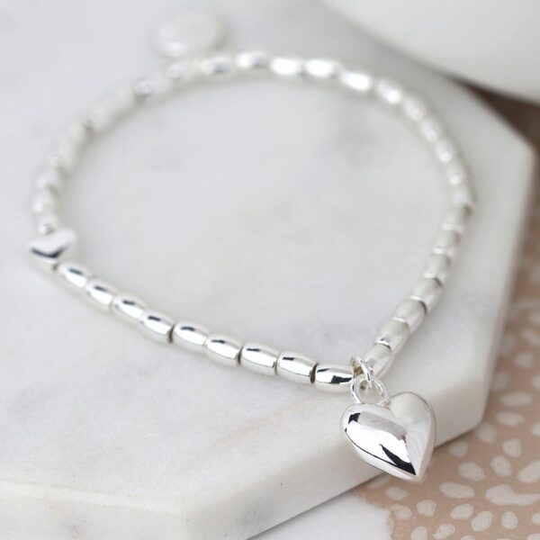 A silver bracelet with a puffed 3d heart charm displayed on a grey marble tray