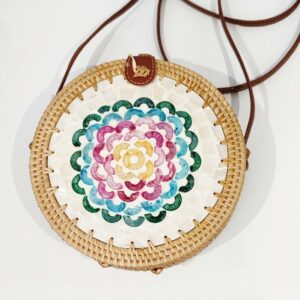 A round handbag with a long strap made of natural rattan with a multi coloured flower decoration on the front made from bits of shell, on a white background