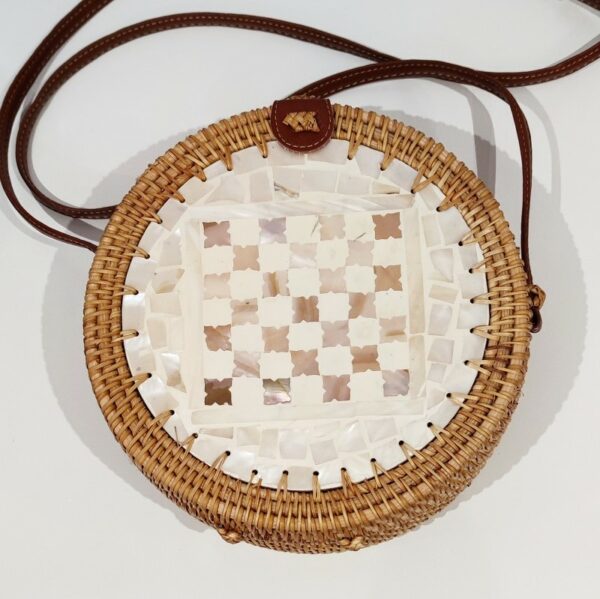 A round handbag with a long brown strap made from natural rattan in the shape of a circle, with natural shell tiles de corating the front, on a white background