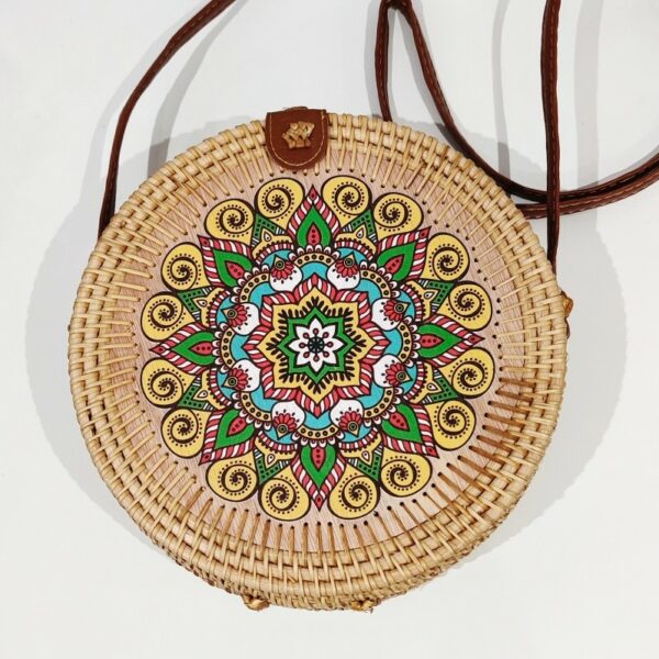 a natural rattan handbag in a circular shape with a long brown strap and a green mandala design on the circular wooden front. On a white background