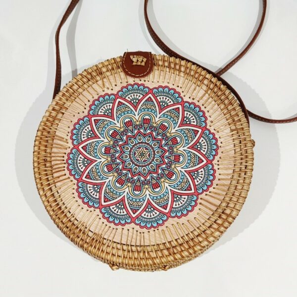 A circular natural rattan handbag, with a long brown handle and a blue mandala design painted on the wooden circular front. On a white background
