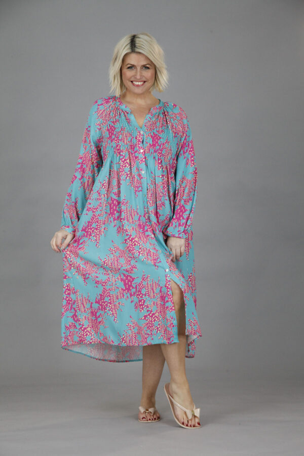A blonde haired lady holding out the sides of a long dress in turquoise with a pink abstract print and long sleeves. She is wearing silver sandals against a grey background