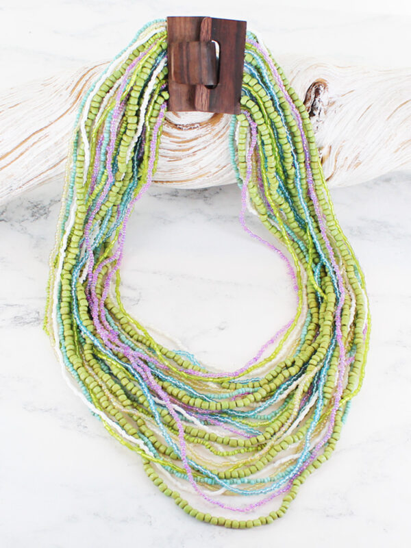 a multi strand necklace in green, blue and lilac with a wooden buckle clasp on a piece of white driftwood