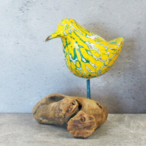 A yellow and green painted wooden carved bird on top of a piece of driftwood against a grey background