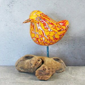 A handcarved bird painted in orange and red standing on a piece of natural coloured driftwood, against a plain grey background