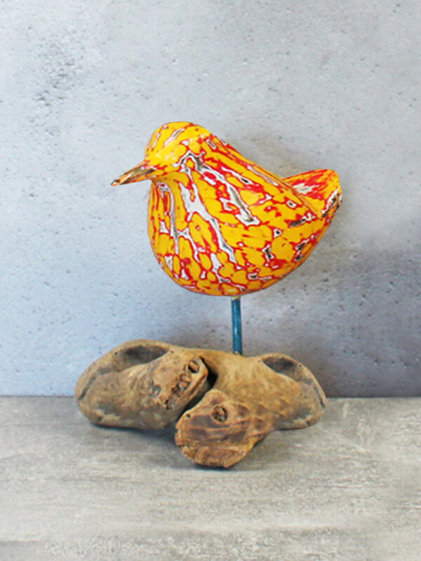 A handcarved bird painted in orange and red standing on a piece of natural coloured driftwood, against a plain grey background