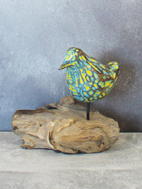 a handpainted carved wooden bird in turquoise blue and yellow, sitting on top of a piece of natural driftwood against a grey background