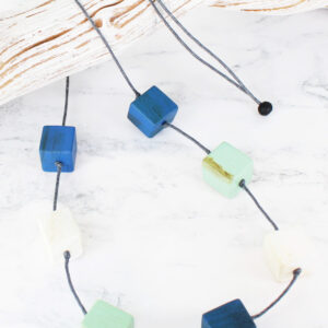 a long necklace made up of painted wooden cubes on a cotton cord in shades of aqua and royal blue, lying across a piece of white driftwood