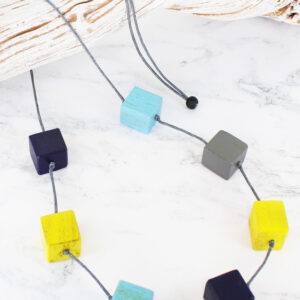a long necklace made of wooden cubes on a rope cord in shades of yellow and blue on a piece of white driftwood