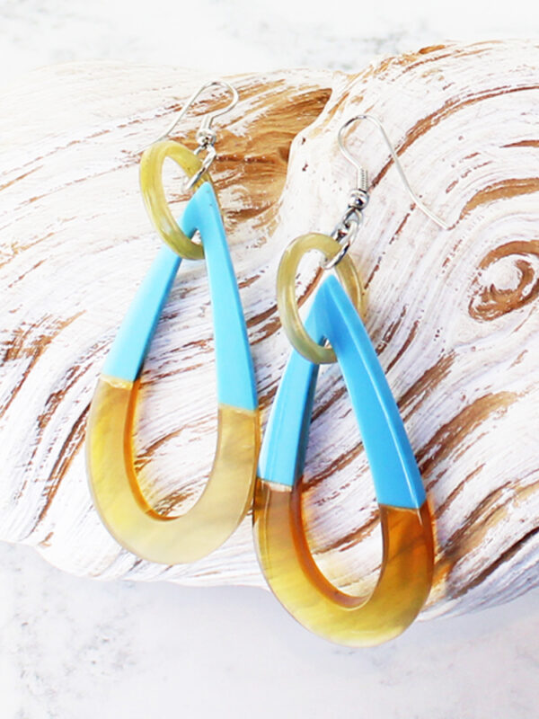 A pair of teardrop shaped earrings made of blue resin and natural horn lying on a piece of white driftwood