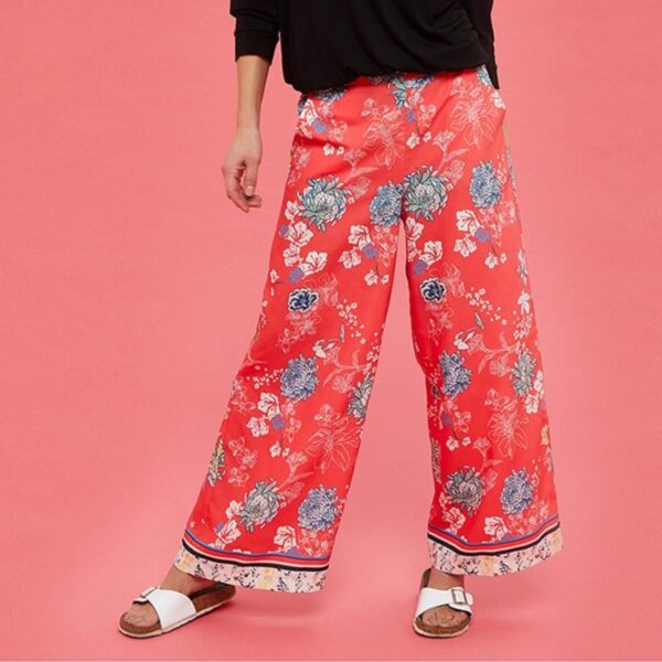 A lady wearing a black sweater and red floral print wide legged trousers and white sandals against a pink background