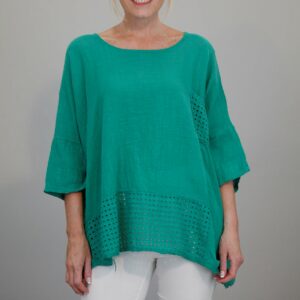 A blonde haired lady in a green cotton 3/4 sleeve top with white trousers on a grey background