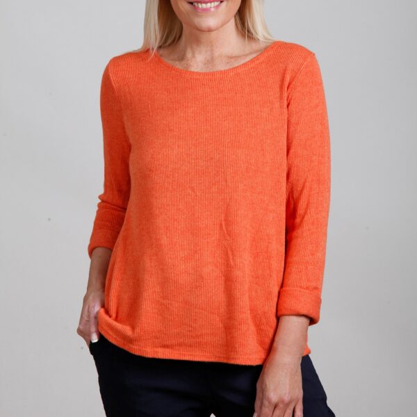 a blonde lady wearing black trousers and an orange top against a buff background