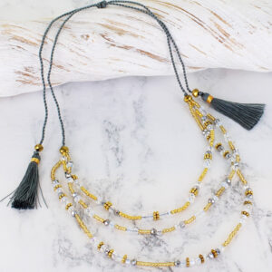 a triple strand crystal bead necklace in shades of grey, gold and clear beads, lying across a pice of white driftwood