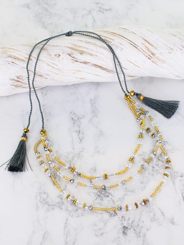 a triple strand crystal bead necklace in shades of grey, gold and clear beads, lying across a pice of white driftwood