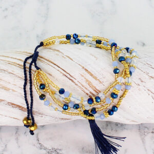 a muti strand gold and blue crystal bead bracelet with a tassle drop, lying on a piece of white driftwood