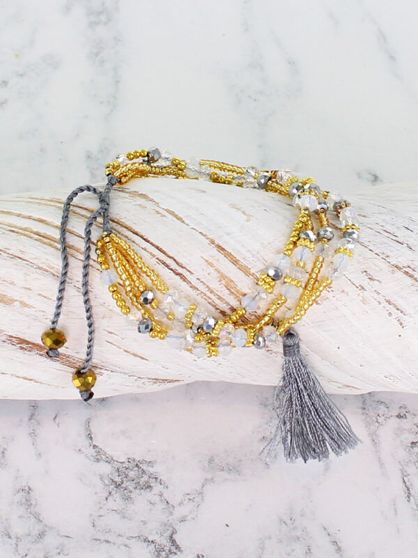 A gold and clear crystal glass bead multi strand bracelet with tie ends, lying on a piece of white driftwood