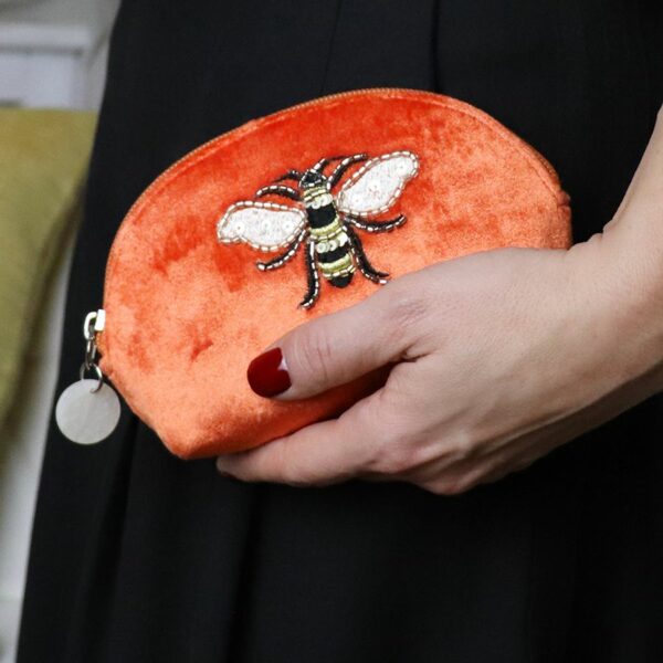 a ladies hand with red nail varnish holding an orange velvet D shaped purse with an embroidered bee on the front