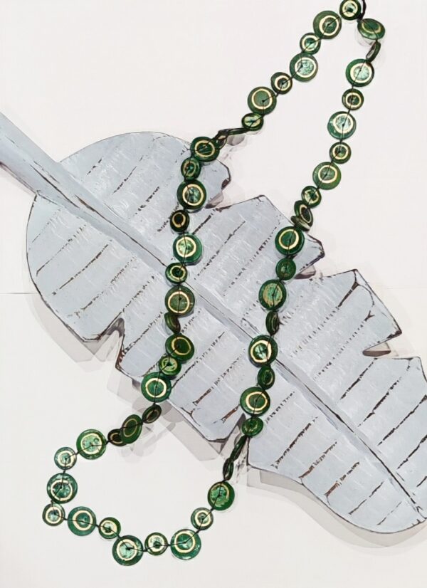 a long necklace made of coconut shell discs in forest green with a silver swirl painted on each disc. It's lying across a pale blue wooden leaf shaped dish