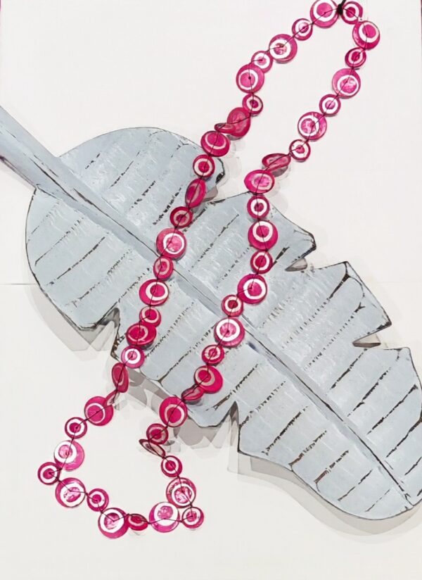 along neclace made up of little coconut shell discs painted in fuschia pink with a silver swirl on each one, laid across a pale blue wooden leaf shaped long dish