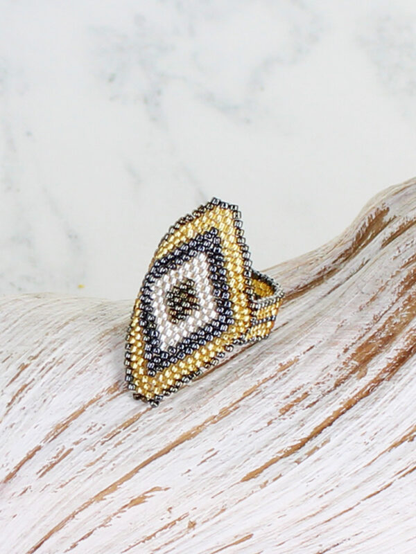 A diamond shaped ring made from glass seed beads in a large diamond design in shades of pewter, gold and silver placed on top of a piece of white driftwood