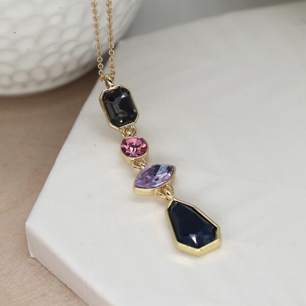 a gold necklace with a multicoloured glass crystal pendant drop, in shades of pink, lilac and dark grey, with a midnight blue drop on the end. it is lying on a white hexagonal tile.
