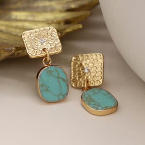 a pair of earrings lying on a white table top. The top part of the earring is an embossed golld metal with an irregular shaped turquoise stone drop hanging from it.