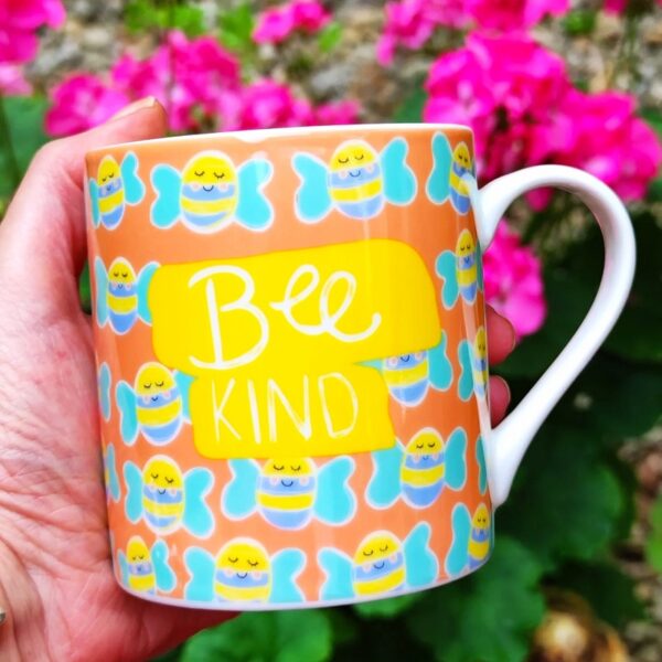 a china mug with a design of blue and yellow bees on it and the slogan "Bee Kind" clasped in a hand against a pink floral background