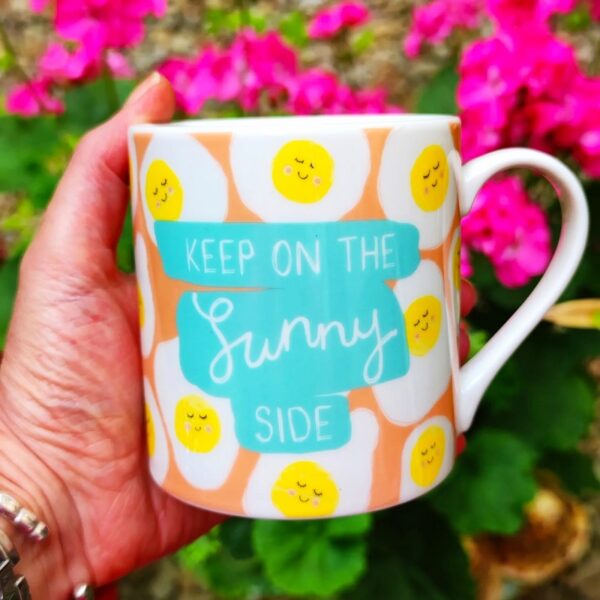 a china mug with an illustration of fried eggs and rthe slogan 'keep on the sunny side' clasped in a hand against a pink flower