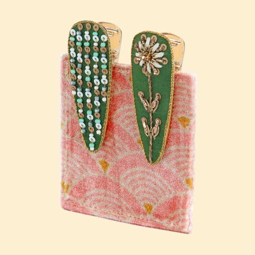 a pair of green jewelled hairclips on a pink velvet card. One is decorated with a gold embroiderd flower and the other in sequin stripes