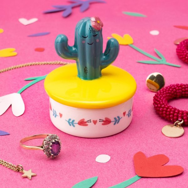 a little ceramic keepsake pot with a yellow lid with a green cactus with a face and flower decoration on the top, sitting on a pink background with some jewellery and coloured confetti around it