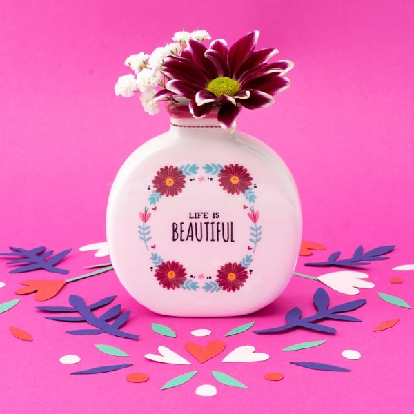 an image of a tiny round bud vase with flowers and the slogan 'life is beautiful' on the front, standing on a pink background with coloured confetti around it, there is a red and white flower in the vase.