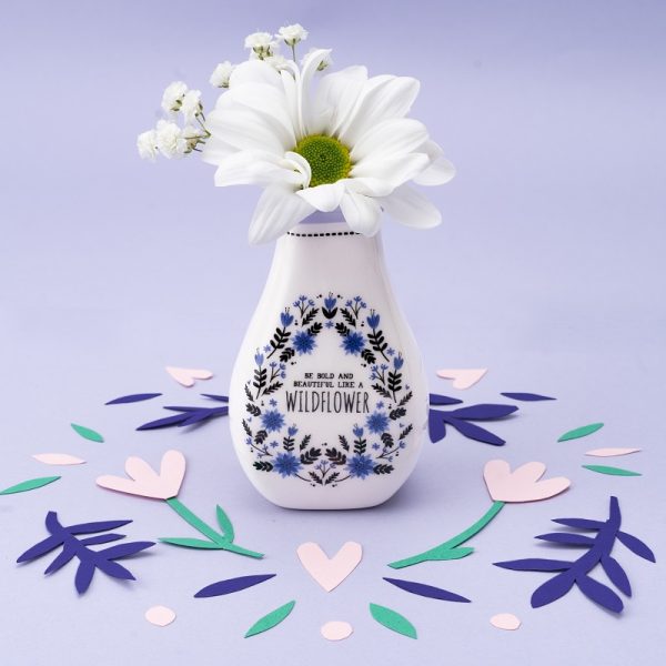 an image of a tiny bud vase with the slogan 'be bold and beautiful like a wildflower' on a lilac background with coloured confetti, there is a white flower in the vase.
