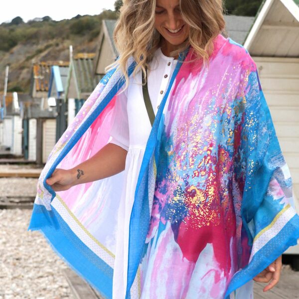 A blonde lady on the beach wearing a scarf wrapped round her shoulders in an abstract print in pink and blue with gold foil overprinting