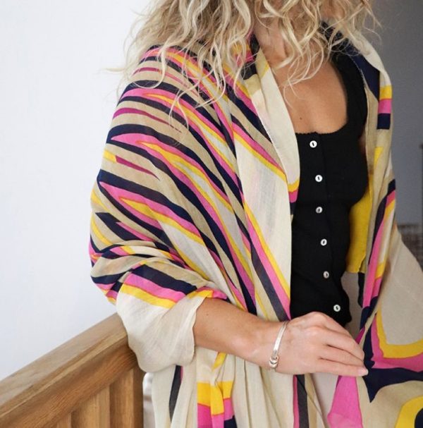 a blonde lady leaning against a wooden bannister with a pink and mustard stripe print scarf over her shoulders