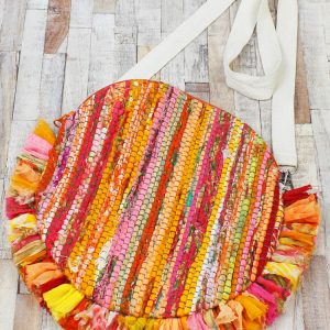A round bag made of strips of recycled sari fabric, with a white webbing cross body strap, and frilled edging around the bottom half of the bag