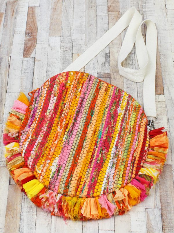 A round bag made of strips of recycled sari fabric, with a white webbing cross body strap, and frilled edging around the bottom half of the bag