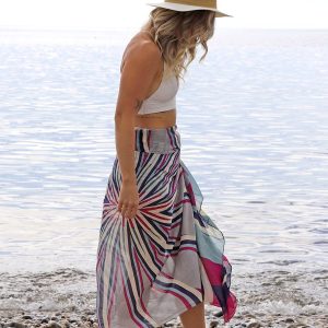 a blonde lady on the beach with the sea behind her, wearing a bikini top and a sarong scarf with a magenta, blue and grey starburst print on it