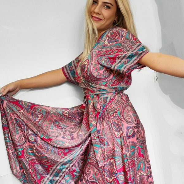 a blonde haired lady wearing a paisley printed long wrap around maxi dress in fuchsia, pink and green .. her arms are outstretched and she is holding the hem of the dress out infront of her.