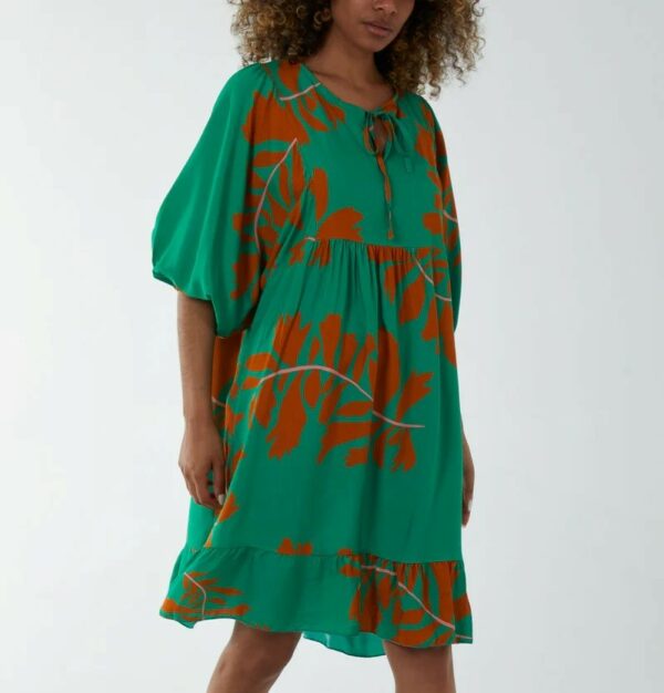 a lady with dark curly hair wearing a knee length smock style dress in jade green with a large leaf print in a rust colour