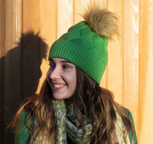 a dark haired girl standing in the sunshine against a slat wood wall, wearing an apple green chevron knit hat with a natural coloured faux fur pompom