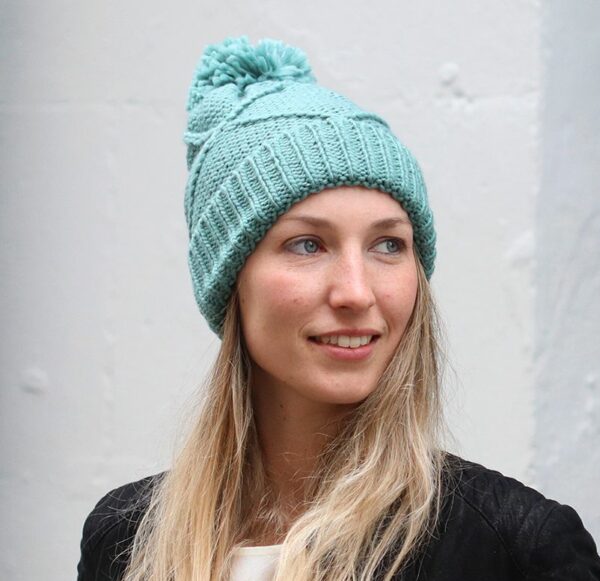 a blonde haired lady standing infront of a white wall, wearing a black jacet, white tee shirt and a cable knit pale teal bobble hat