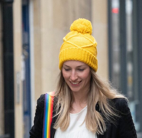 a blonde haired lady wearing a navy jacket and white tee shirt, she has a rainbow strap over her shoulder and a golden yellow cable knit bobble hat on her head