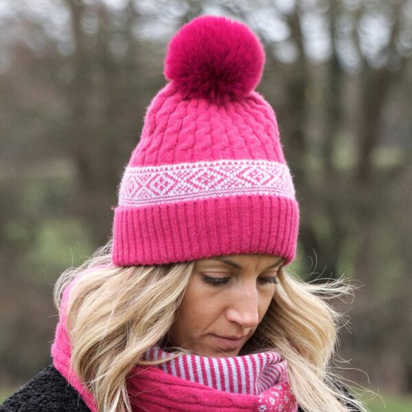 a blonde lady outside, wearing a pink cable knit bobble hat with a large pink faux fur pompom on the top