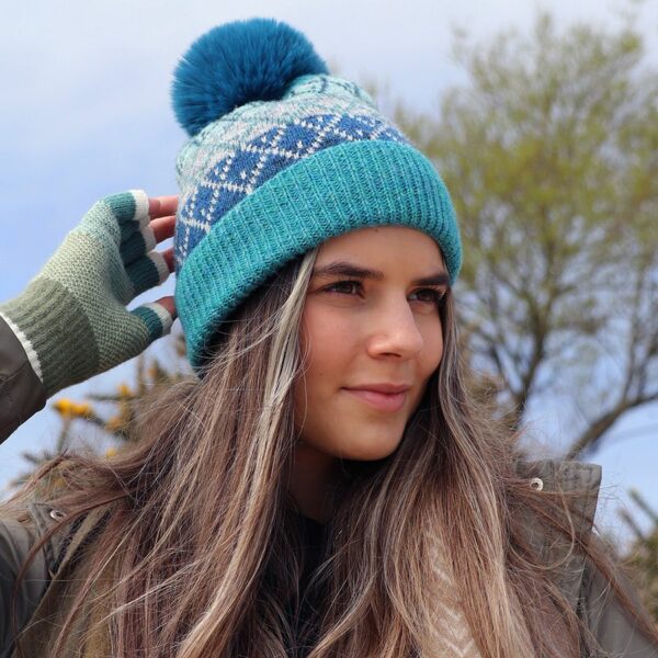 a girl with long hair outside with a tree in the background, she is wearing a teal and grey knitted hat with a large teal faux fur pompom on the top