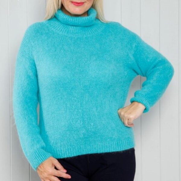 a blonde lady against a white wood background wearing navy trousers and a fluffy knit turquoise roll neck jumper