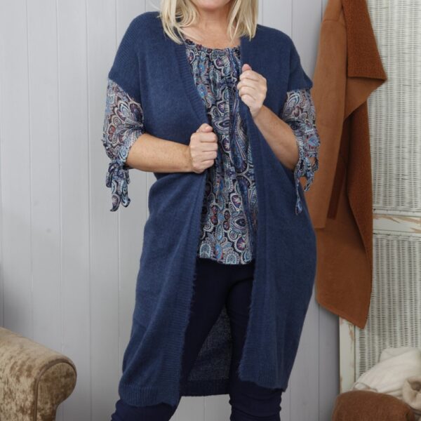 a blonde lady wearing a blue silk paisley print blouse underneath a sleeveless long knit open front cardigan